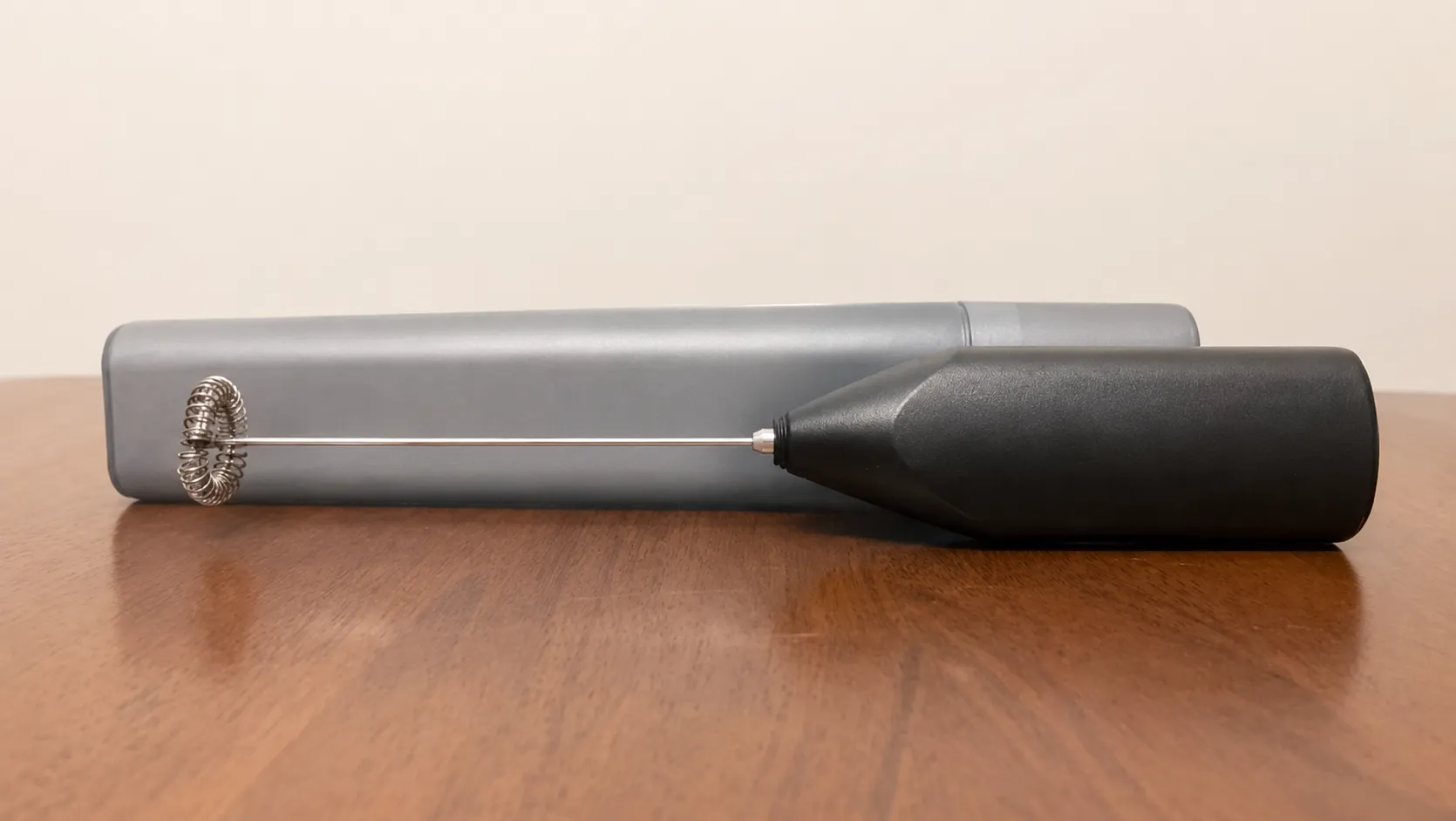 Original supplier-style product photo of a portable electric milk frother on a wooden table with a beige wall background